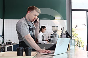 Young handsom male barista wearing apron, talking order on mobile phone and laptop computer while standing behind coffee shop