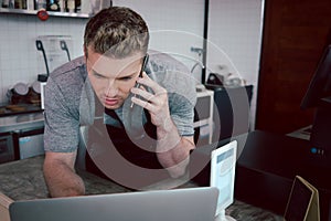 Young handsom male barista wearing apron, talking order on mobile phone and laptop computer while standing behind coffee shop