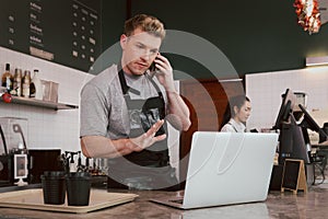 Young handsom male barista wearing apron, talking order on mobile phone and laptop computer while standing behind coffee shop