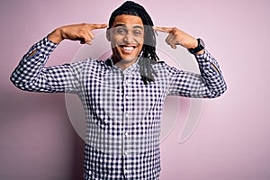 Young handsome african american afro man with dreadlocks wearing casual shirt smiling pointing to head with both hands finger,