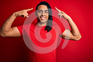 Young handsome african american afro man with dreadlocks wearing red casual t-shirt smiling pointing to head with both hands