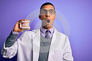 Young handsome african american dentist man holding denture teeth over purple background scared in shock with a surprise face,