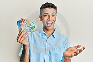 Young handsome african american man holding australian dollars celebrating achievement with happy smile and winner expression with