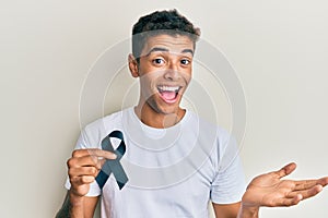 Young handsome african american man holding black ribbon celebrating achievement with happy smile and winner expression with
