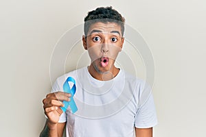 Young handsome african american man holding blue ribbon scared and amazed with open mouth for surprise, disbelief face