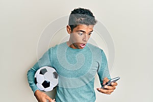 Young handsome african american man holding football ball looking at smartphone afraid and shocked with surprise and amazed