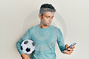 Young handsome african american man holding football ball looking at smartphone relaxed with serious expression on face