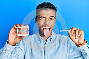 Young handsome african american man holding invisible aligner orthodontic and braces sticking tongue out happy with funny