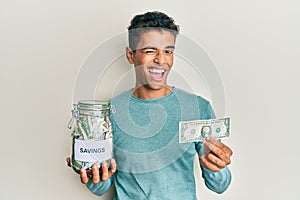 Young handsome african american man holding jar with savings winking looking at the camera with sexy expression, cheerful and