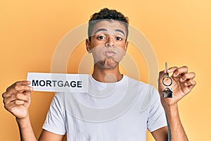 Young handsome african american man holding paper with mortgage word and house keys puffing cheeks with funny face