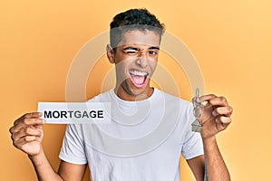 Young handsome african american man holding paper with mortgage word and house keys winking looking at the camera with sexy