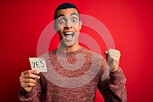 Young handsome african american man holding reminder paper with yes message screaming proud and celebrating victory and success