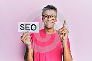 Young handsome african american man holding seo message paper smiling with an idea or question pointing finger with happy face,