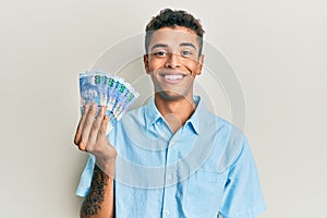 Young handsome african american man holding south african 100 rand banknotes looking positive and happy standing and smiling with