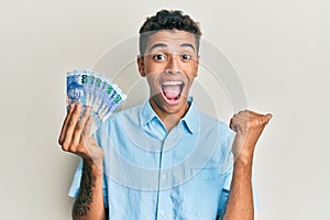 Young handsome african american man holding south african 100 rand banknotes pointing thumb up to the side smiling happy with open
