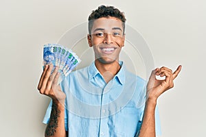 Young handsome african american man holding south african 100 rand banknotes smiling happy pointing with hand and finger to the