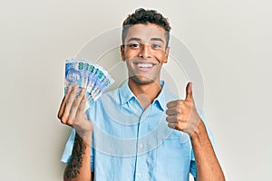 Young handsome african american man holding south african 100 rand banknotes smiling happy and positive, thumb up doing excellent