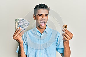 Young handsome african american man holding virtual currency bitcoin and dollars winking looking at the camera with sexy