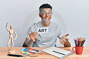Young handsome african american man painter sitting at art studio holding art word celebrating achievement with happy smile and