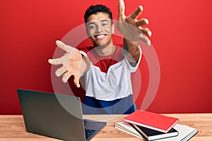 Young handsome african american man studying for school using laptop looking at the camera smiling with open arms for hug