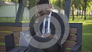 Young handsome African American man in suit putting on headphones and listening to music in sunny summer park. Portrait