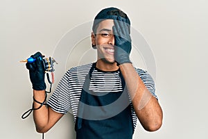 Young handsome african american man tattoo artist wearing professional uniform and gloves holding tattooer machine covering one