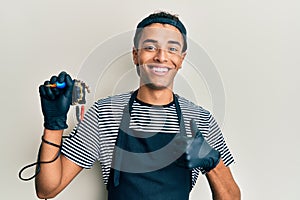 Young handsome african american man tattoo artist wearing professional uniform and gloves holding tattooer machine doing happy