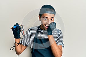 Young handsome african american man tattoo artist wearing professional uniform and gloves holding tattooer machine laughing and