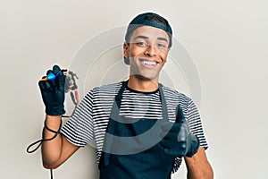 Young handsome african american man tattoo artist wearing professional uniform and gloves holding tattooer machine pointing