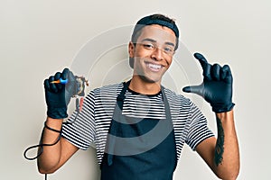 Young handsome african american man tattoo artist wearing professional uniform and gloves holding tattooer machine smiling and