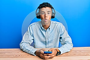 Young handsome african american man using smartphone wearing headphones making fish face with mouth and squinting eyes, crazy and