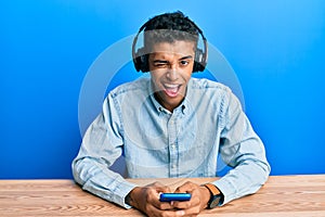 Young handsome african american man using smartphone wearing headphones winking looking at the camera with sexy expression,