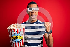 Young handsome african american man watching movie using 3d glasses eating popcorns angry and mad raising fist frustrated and