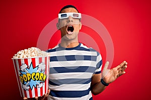 Young handsome african american man watching movie using 3d glasses eating popcorns crazy and mad shouting and yelling with