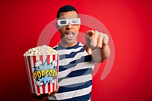 Young handsome african american man watching movie using 3d glasses eating popcorns pointing displeased and frustrated to the