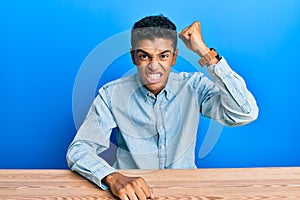Young handsome african american man wearing casual clothes sitting on the table angry and mad raising fist frustrated and furious
