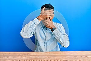 Young handsome african american man wearing casual clothes sitting on the table covering eyes and mouth with hands, surprised and