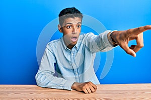 Young handsome african american man wearing casual clothes sitting on the table pointing with finger surprised ahead, open mouth