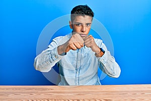 Young handsome african american man wearing casual clothes sitting on the table punching fist to fight, aggressive and angry