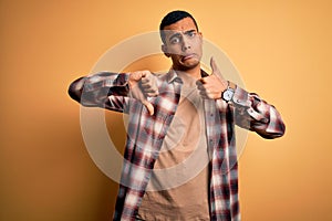 Young handsome african american man wearing casual shirt standing over yellow background Doing thumbs up and down, disagreement