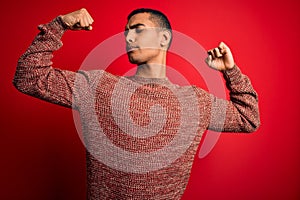 Young handsome african american man wearing casual sweater standing over red background showing arms muscles smiling proud