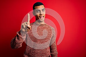 Young handsome african american man wearing casual sweater standing over red background showing and pointing up with finger number