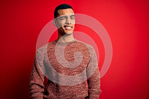 Young handsome african american man wearing casual sweater standing over red background winking looking at the camera with sexy