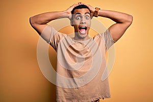 Young handsome african american man wearing casual t-shirt standing over yellow background Crazy and scared with hands on head,
