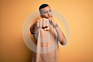 Young handsome african american man wearing casual t-shirt standing over yellow background Punching fist to fight, aggressive and