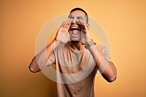 Young handsome african american man wearing casual t-shirt standing over yellow background Shouting angry out loud with hands over