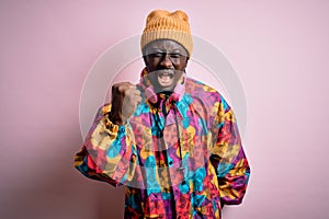 Young handsome african american man wearing colorful coat and cap over pink background angry and mad raising fist frustrated and