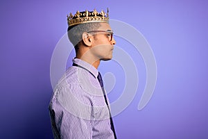 Young handsome african american man wearing golden crown of king over purple background looking to side, relax profile pose with