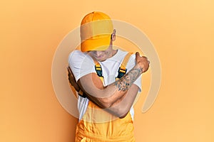 Young handsome african american man wearing handyman uniform over yellow background hugging oneself happy and positive, smiling