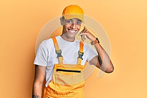 Young handsome african american man wearing handyman uniform over yellow background smiling doing phone gesture with hand and